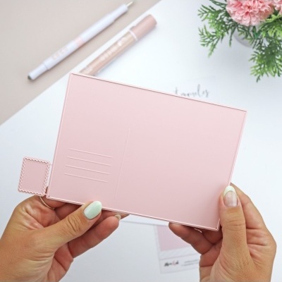 Estojos rosa claro com detalhes quadrados segurados por mãos sobre mesa branca com flores e materiais de papel