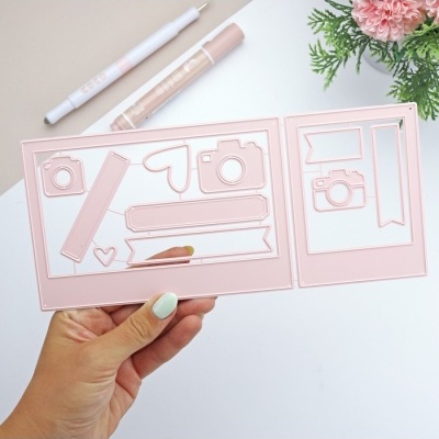 Cortantes de papel rosa claro com formas diversas segurados por mão com unhas pintadas, sobre fundo branco com caneta e flores ao lado.