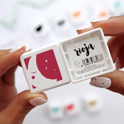 Produto cosmético quadrado branco com desenho vermelho e rótulo 'rioja' segurado por mãos com unhas pintadas