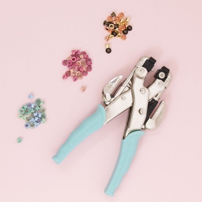 Alicate para ilhós cromado com pegas azuis claras com ilhós coloridos sobre fundo rosa