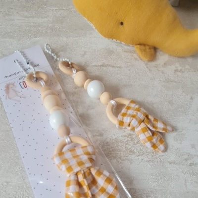 Acessórios de bebé em madeira, silicone e tecido com padrão xadrez amarelo e branco, um embalado e outro fora do saco junto a um brinquedo de pelúcia amarelo