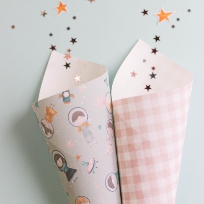Dois cones de papel decorativo, um cinza com bonecos astronautas e outro rosa quadriculado, com confetis em estrelas a sair deles.