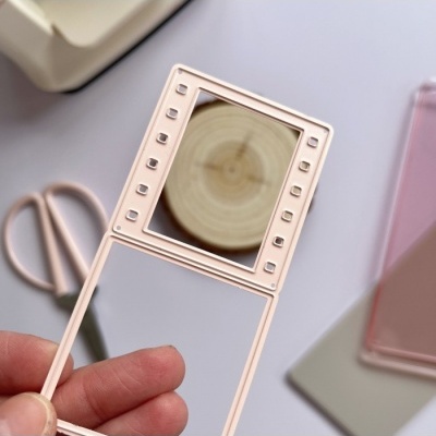 Moldura rosa pálido com espelho quadrado e recortes, segurada por mão, fundo com tesoura e capas de telemóvel