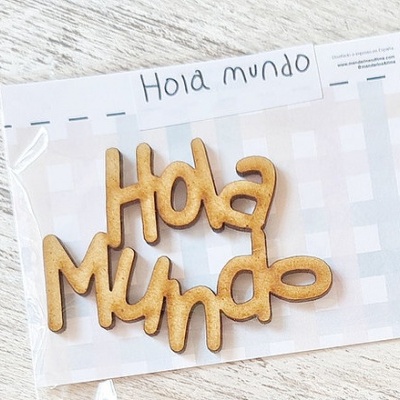 Decoração em madeira com texto 'Hola Mundo' em embalagem transparente sobre fundo branco quadriculado