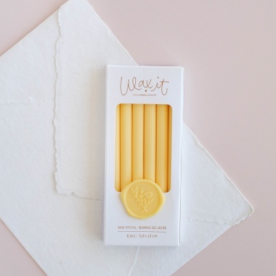 Varetas de cera amarelas para lacrar em caixa branca com selo amarelo sobre papel branco.