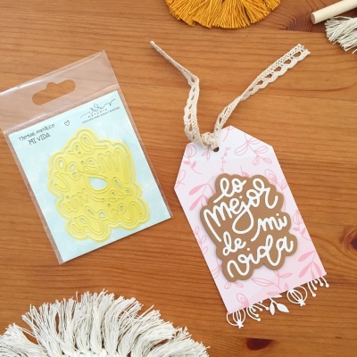 Dois produtos sobre mesa de madeira: estêncil amarelo em embalagem e etiqueta de papel rosa com frase em espanhol