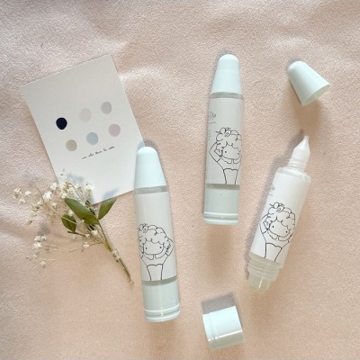 Frascos brancos com tampa azul clara sobre tecido rosa claro, com ilustração de rosto infantil e cartão de cores pastel.