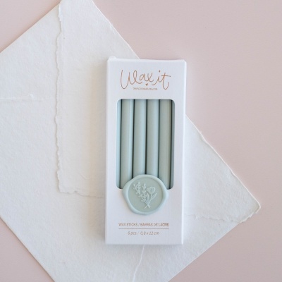 Conjunto de varetas de cera de lacre cinza claro em caixa branca com selo decorativo sobre papel branco e fundo rosa