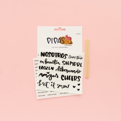 Pacote de autocolantes com frases manuscritas em português e espanhol preto sobre fundo branco e palito de madeira em fundo rosa.