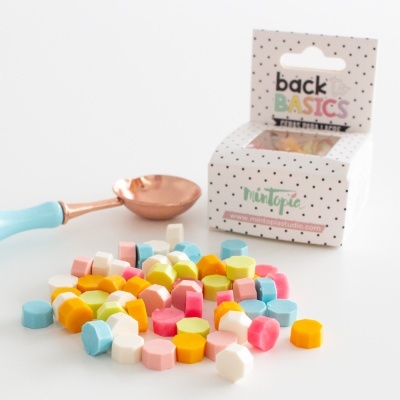 Caixa de ceras coloridas e colher metálica cor de cobre com cabo azul claro sobre superfície branca