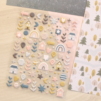 Autocolantes em 3D coloridos em tons pastel sobre mesa de madeira com papel decorativo ao lado