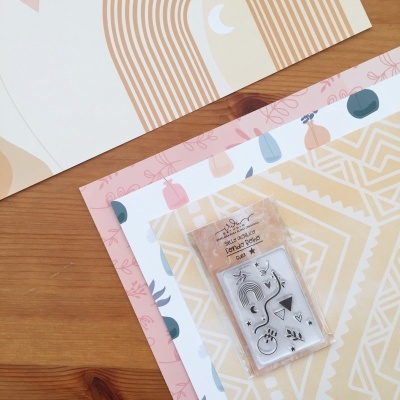 Folhas de papel decorativo em tons pastel com carimbos de silicone embalados em plástico sobre madeira.