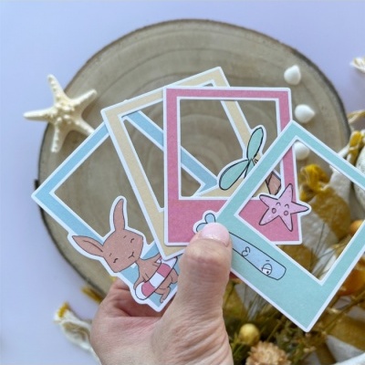 Quatro molduras de cartão coloridas com desenhos infantis seguradas por uma mão sobre fundo com prato de madeira e conchas