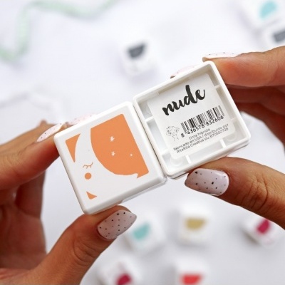 Estojo branco aberto com tom nude e etiqueta 'nude' segurado por mãos com unhas pintadas