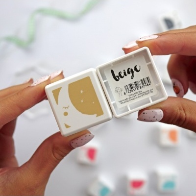 Caixa pequena branca com desenho bege e etiqueta 'beige', mãos segurando com unhas pintadas