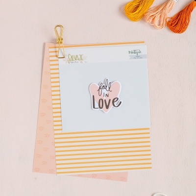 Cadernos com adesivo 'fall IN Love', novelos de linha coloridos, tesoura branca e prato decorativo sobre superfície rosa clara