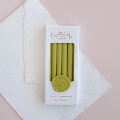 Pacote branco com seis varetas de cera verde-oliva e selo redondo verde, em fundo claro com papel e superfície rosa