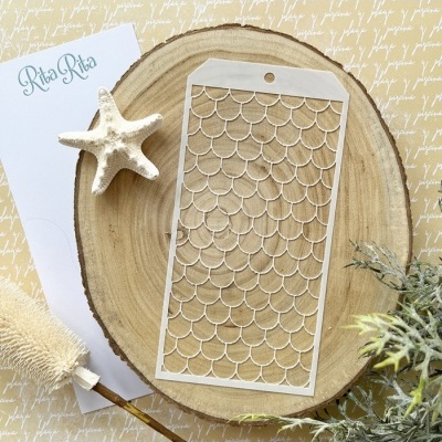 Stencil de plástico branco com padrão de escamas sobre madeira redonda com elementos decorativos e papel com texto Rita Rita.