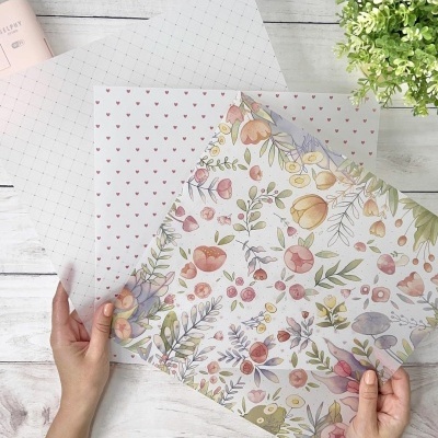 Folhas de papel de embrulho com padrões de corações e flores coloridas, seguradas por mãos sobre mesa branca texturizada