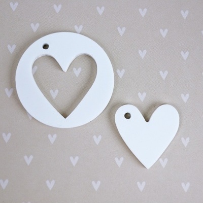Dois pendentes brancos em formato redondo com recorte de coração e em forma de coração sobre fundo bege com padrão de corações.