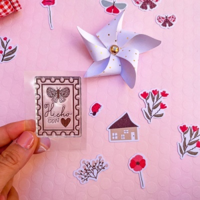 Adesivo quadrado transparente com borboleta e texto, rodeado por recortes de papel e um pinwheel branco.