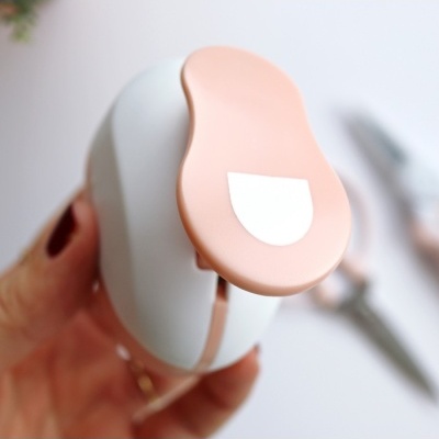 Ferramenta de papelaria branca com parte rosa claro e etiqueta branca, em fundo desfocado com outras ferramentas.