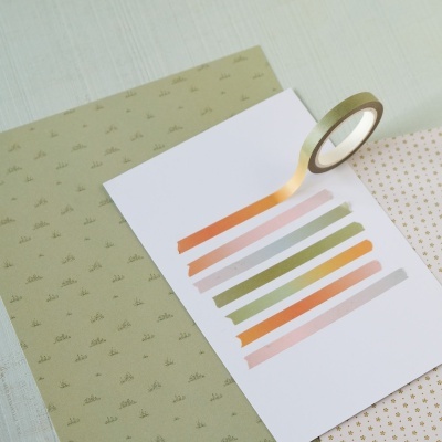 Folha branca com tiras de fita adesiva colorida e rolo de fita adesiva ao lado sobre tecidos com padrões pequenos