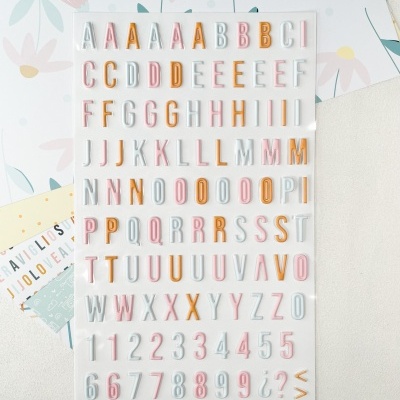 Folha com letras adesivas coloridas em tons pastel colocada sobre mesa clara com papéis decorativos florais