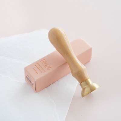 selo de lacre com cabo de madeira e base metálica dourada próximo de uma caixa rosa e papel branco