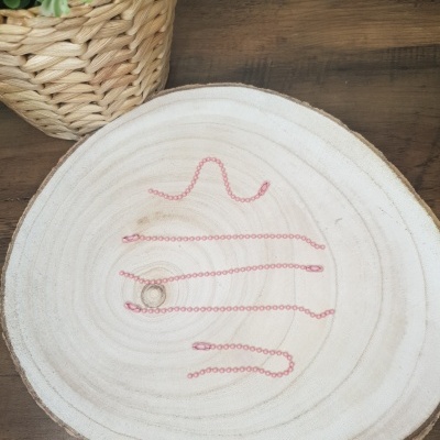 Colar com corrente rosa claro e pendente dourado numa base de madeira clara com planta em vaso ao fundo