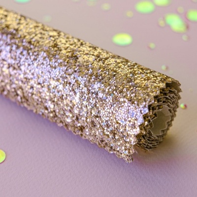 Rolo de tecido decorativo dourado e prateado com textura glitter sobre superfície rosa com confetes coloridos