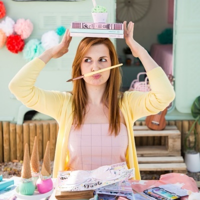 Mulher ruiva com casaco amarelo segura livros na cabeça e pincel na boca num espaço decorado com cones de gelado e utensílios de pintura.