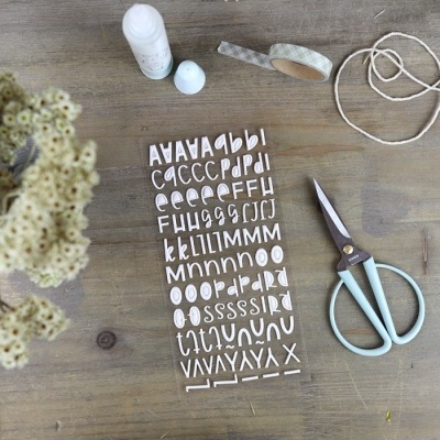 folha de letras autocolantes brancas com tesoura azul, fita adesiva, cordão branco e frasco branco sobre mesa de madeira com flores secas