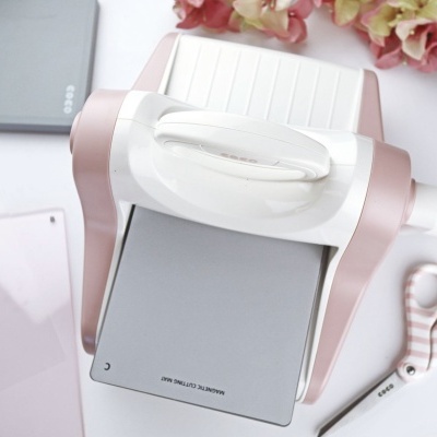 Máquina de corte manual branca e rosa claro com placa cinzenta e acessórios ao redor