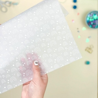Folha translúcida com padrão branco de arcos-íris e estrelas segurada por uma mão sobre fundo bege com objetos coloridos desfocados.