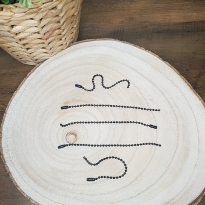 Vários cordões esferas pretas metálicas sobre tábua de madeira clara, vaso de palha com plantas ao lado