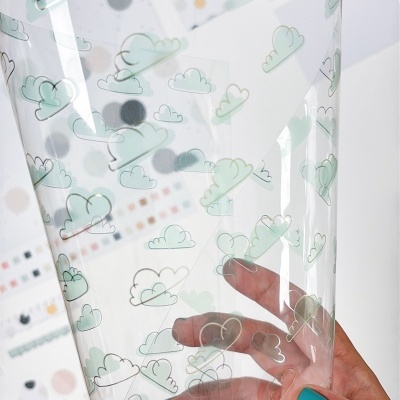 Saco plástico transparente com nuvens azuis segurado por mão com unhas azuis, fundo com adesivos