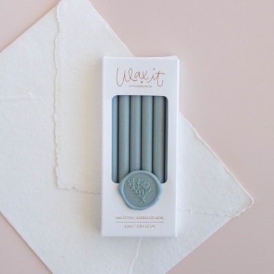 Caixa branca com velas finas azuis e selo azul sobre papel branco texturado