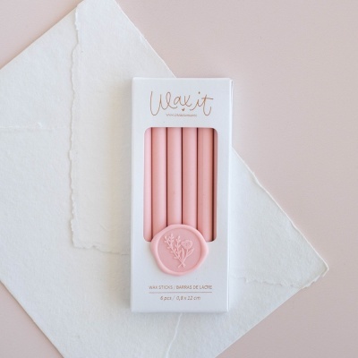 Caixa branca com seis bastões de cera rosa e selo floral, sobre papel e fundo rosa