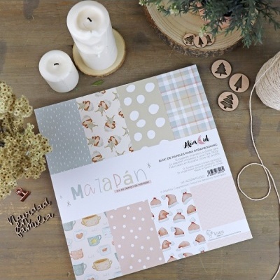 Bloco de papel para scrapbook com diversos padrões decorativos em superfície de madeira com velas, flores secas e acessórios de artesanato.