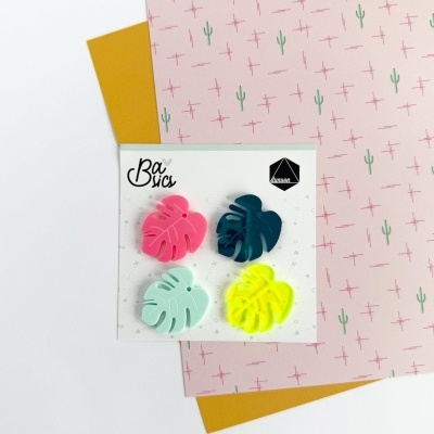 Clipes de papel coloridos em forma de folha de monstera sobre fundo com papéis decorativos.