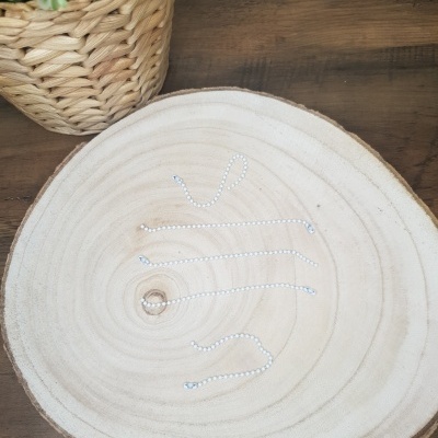 Anel dourado sobre tábua redonda de madeira clara junto a vaso de cesto com plantas, tudo sobre mesa de madeira escura