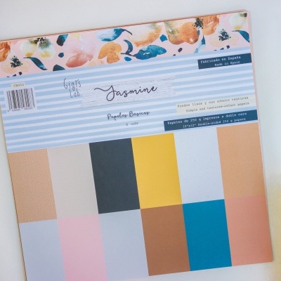 Folhas de papel para scrapbooking coloridas com etiqueta da marca Jasmine sobre faixa azul e branca, em fundo branco com flores e decoração natural