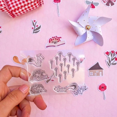 Conjunto de carimbos transparentes com desenhos de esquilos e flores, mão segurando, fundo rosa com decoração floral e roda de moinho.