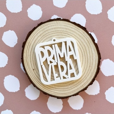 Objeto decorativo branco com palavra PRIMAVERA sobre base de madeira e fundo cor de rosa com pontos brancos
