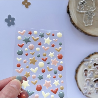 Folha de autocolantes tridimensionais coloridos e brilhantes com formas de estrelas e corações, segurada por uma mão.