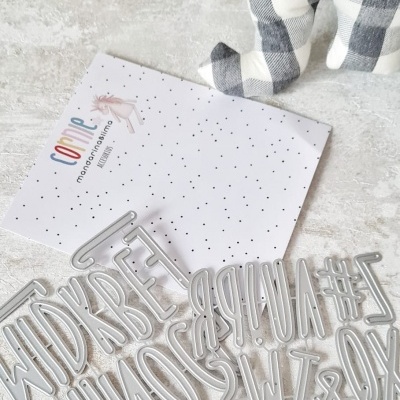 Moldes metálicos de letras e símbolos sobre superfície branca, cartão com texto e peluche xadrez cinza e branco ao fundo