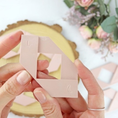 Mãos segurando objeto quadrado rosa claro com símbolos, flores e base de madeira ao fundo