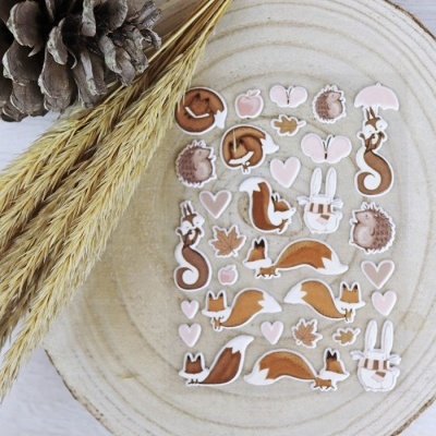 Autocolantes ilustrados de animais e elementos da natureza em tons castanhos e rosa sobre madeira clara com espigas secas ao lado.