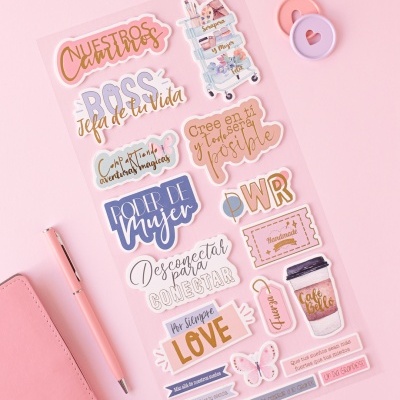 Folha de autocolantes coloridos com textos motivacionais em espanhol e imagens de chá, café e flores sobre fundo rosa com caneta e caderno rosa.